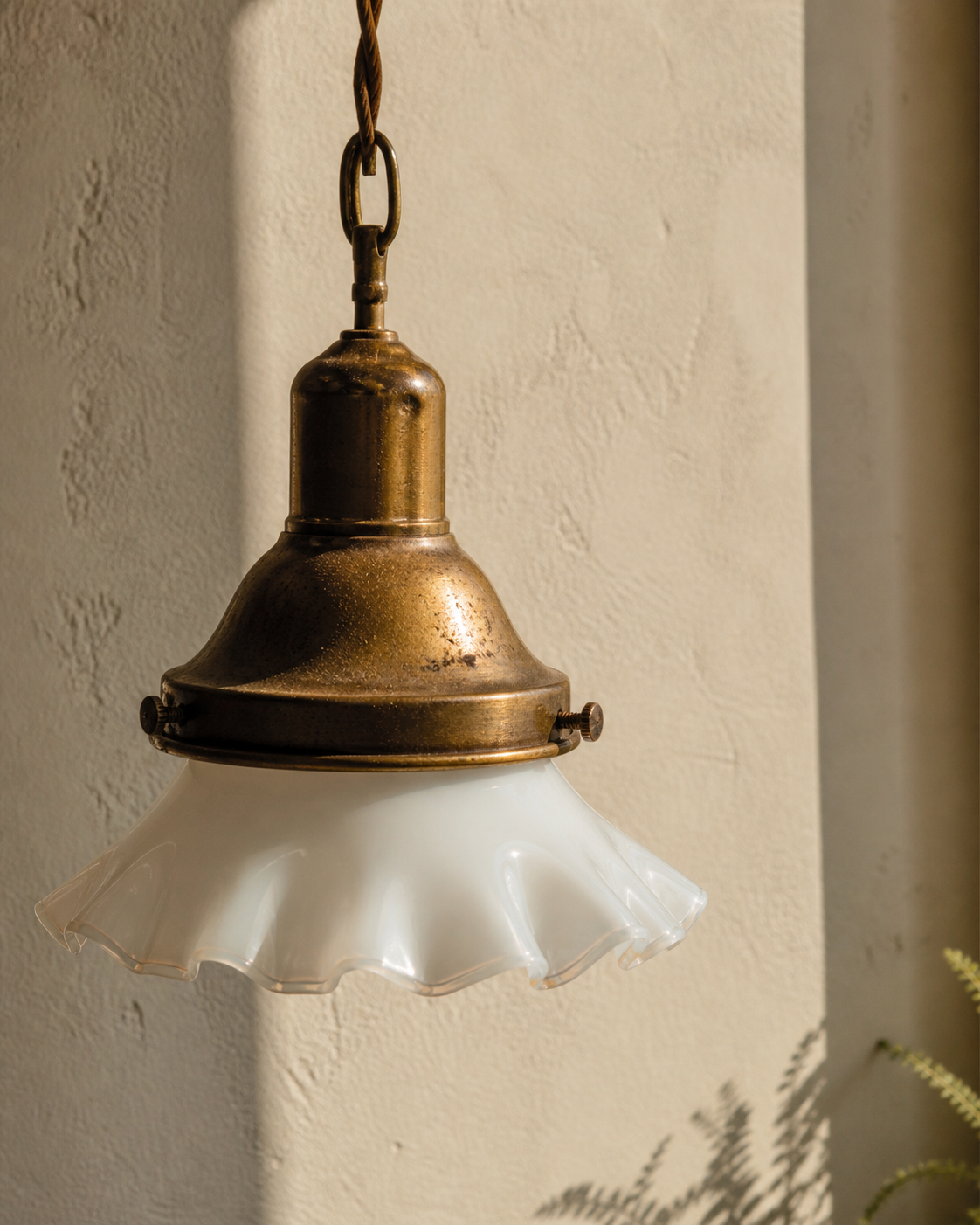 Curateur Atelier — an antique brass pendant lamp with a fluted milk-glass shade against a soft plaster wall